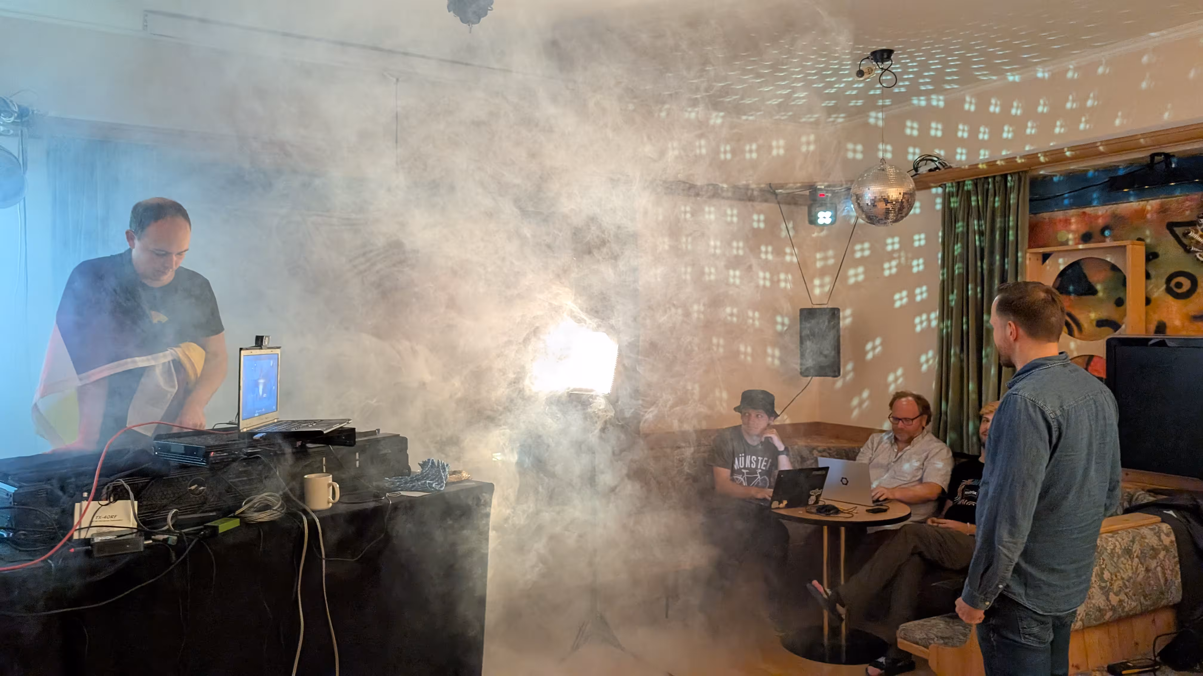 Someone is standing at the DJ desk, wearing a pride flag, the room is filled with disco fog. More people are sitting on their laptops at a round table.